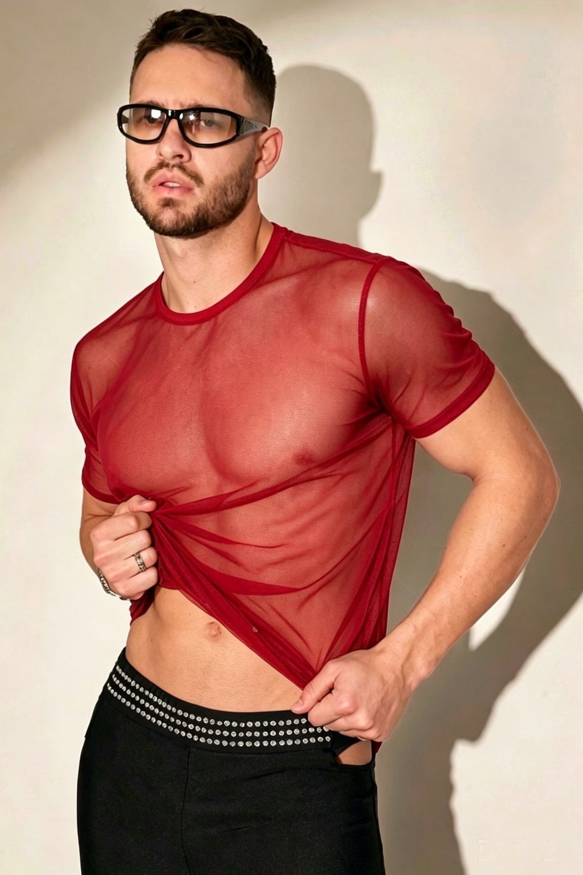A man wearing a sheer red short-sleeve mesh shirt and black pants with a silver studded waistband, posing against a beige wall with a strong shadow.