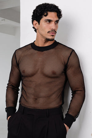 A man wearing a black long-sleeve fishnet mesh shirt and black trousers, standing against a white wall.