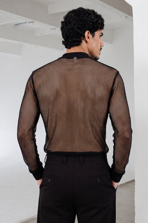 A man viewed from behind wearing a black long-sleeve mesh shirt and black trousers in a minimalist white room.