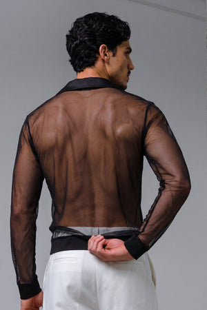 A man shown from the back wearing a sheer black long-sleeve button-up shirt over white trousers, standing against a gray studio background with a concrete-textured column on the right.