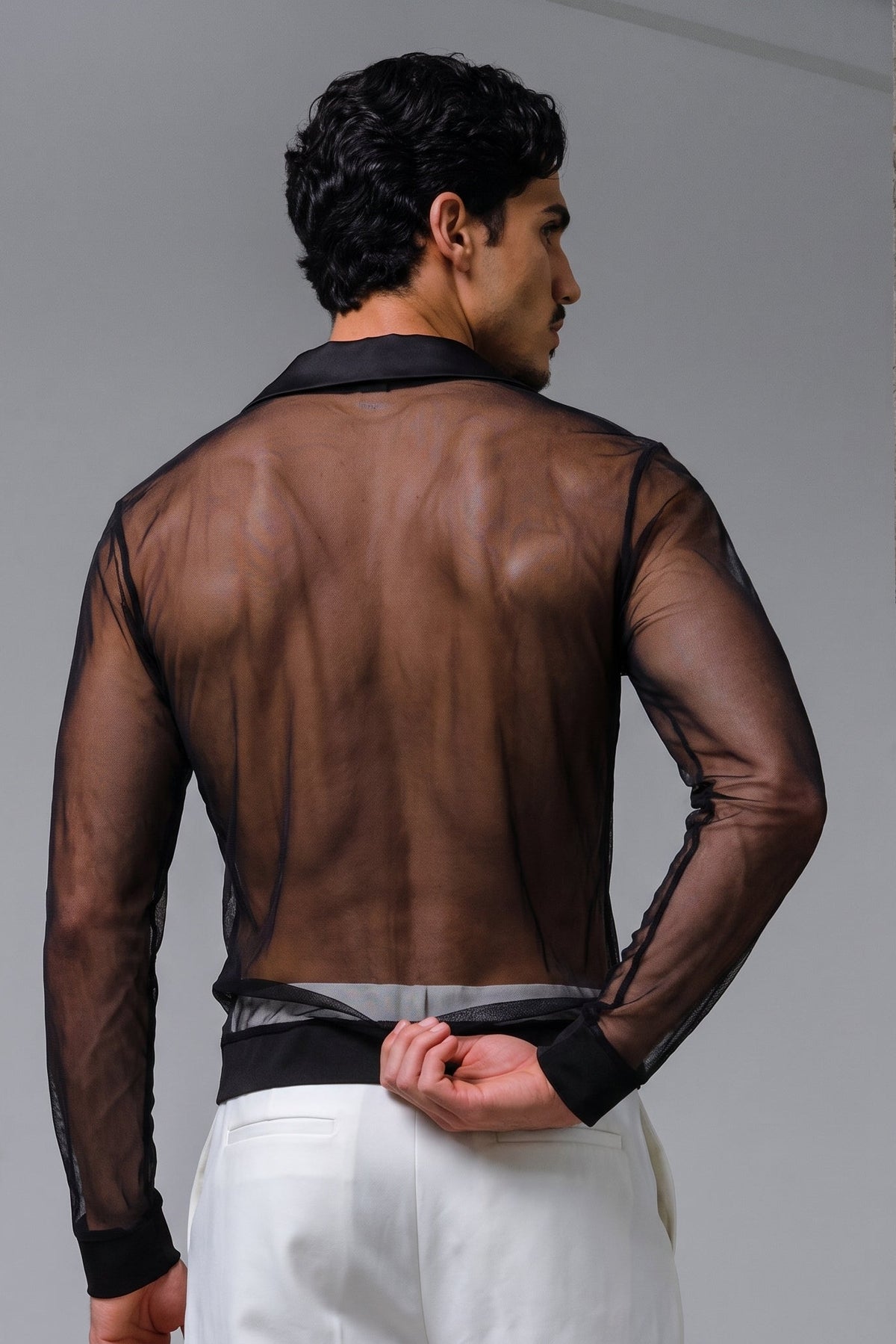 A man shown from the back wearing a sheer black long-sleeve button-up shirt over white trousers, standing against a gray studio background with a concrete-textured column on the right.