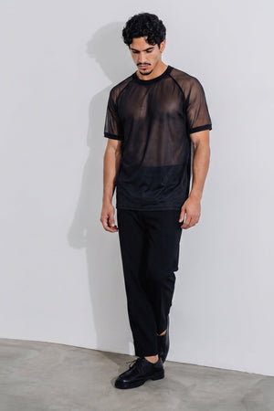 A man wearing a black short-sleeve mesh t-shirt and black pants, standing against a light gray wall.