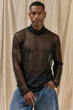 A man wearing a sheer black long-sleeve mock neck t-shirt and blue jeans, standing against a beige draped fabric backdrop.