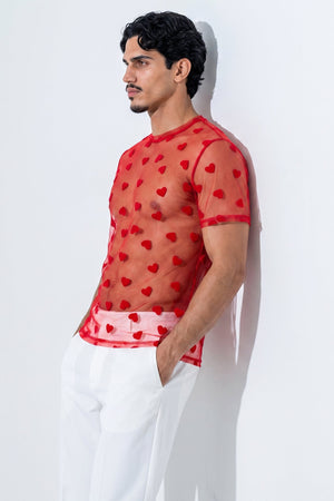 A man wearing a sheer red short-sleeve mesh top with a red heart pattern, paired with white trousers, leaning against a white wall.