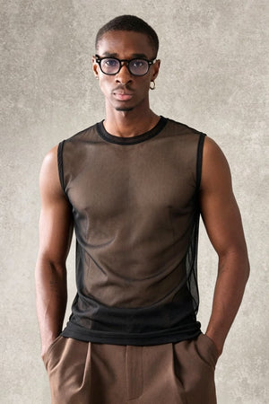 A man wearing a sheer black mesh sleeveless top over a brown undershirt, paired with brown trousers, standing against a textured gray background.