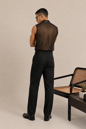 A man stands with his back to the camera wearing a sleeveless black mesh shirt, black trousers, and black dress shoes in a studio setting with a woven chair and side table.