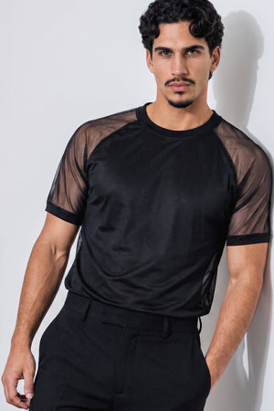 A man wearing a black short-sleeve athletic mesh shirt with sheer shoulder panels and black trousers, standing against a light gray wall.