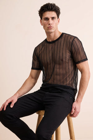 A man wearing a sheer black short-sleeve mesh t-shirt with vertical stripes and black pants, sitting on a wooden stool against a solid beige background.