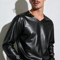 A man wearing a black faux-leather long-sleeve V-neck shirt, aviator sunglasses, and light-wash jeans, standing against a white wall with a shadow.
