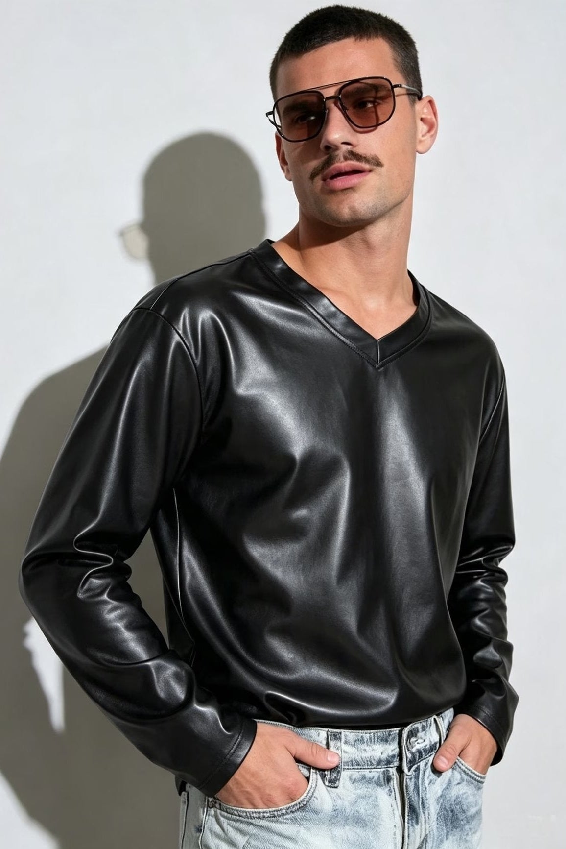 A man wearing a black faux-leather long-sleeve V-neck shirt, aviator sunglasses, and light-wash jeans, standing against a white wall with a shadow.