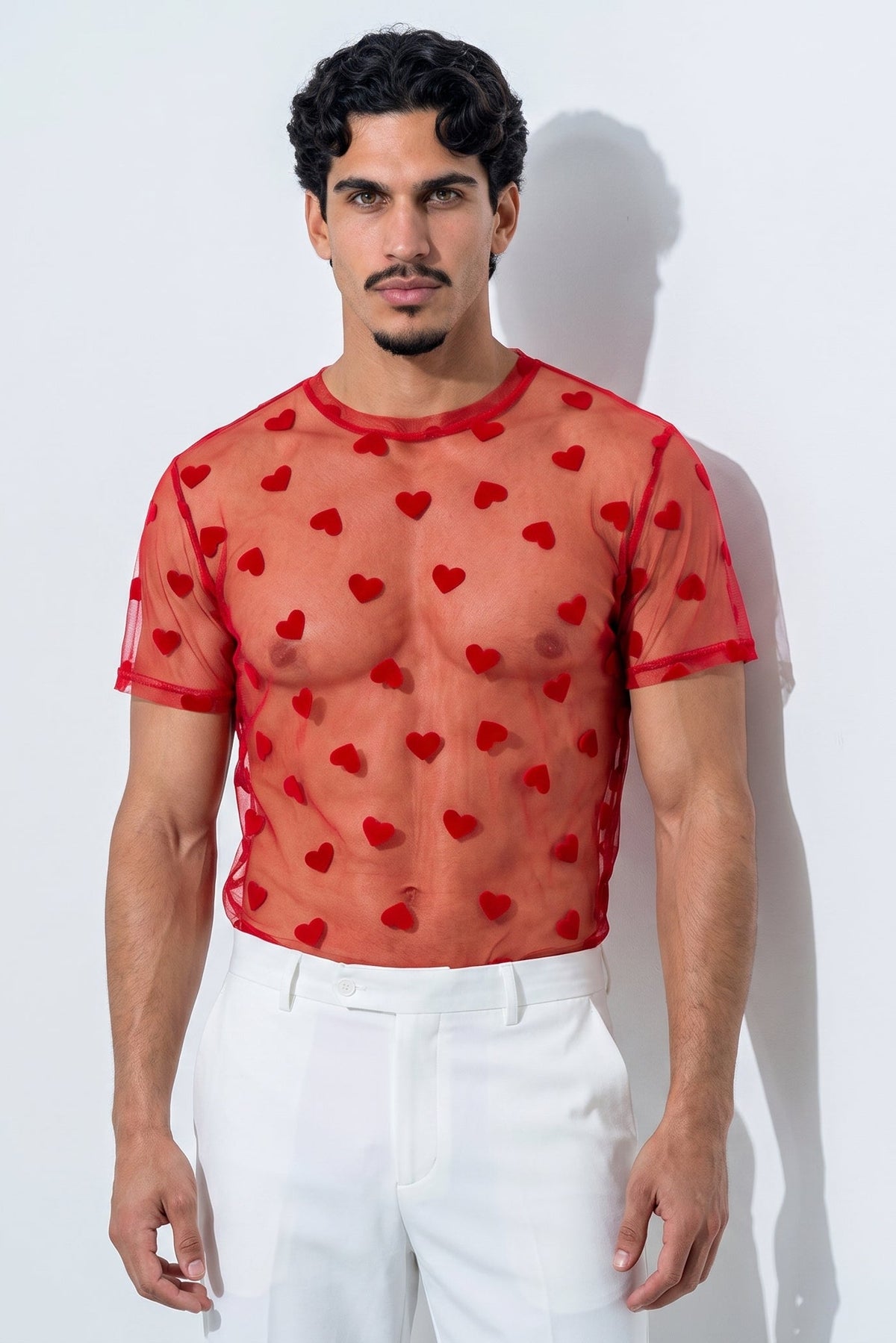 A man modeling a sheer red short-sleeve mesh top with an allover red heart pattern, paired with white pants, standing against a light gray wall.
