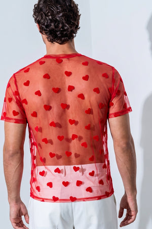 A man viewed from behind wearing a sheer red short-sleeve top covered in red heart motifs, paired with white pants, against a light gray wall with a soft shadow.