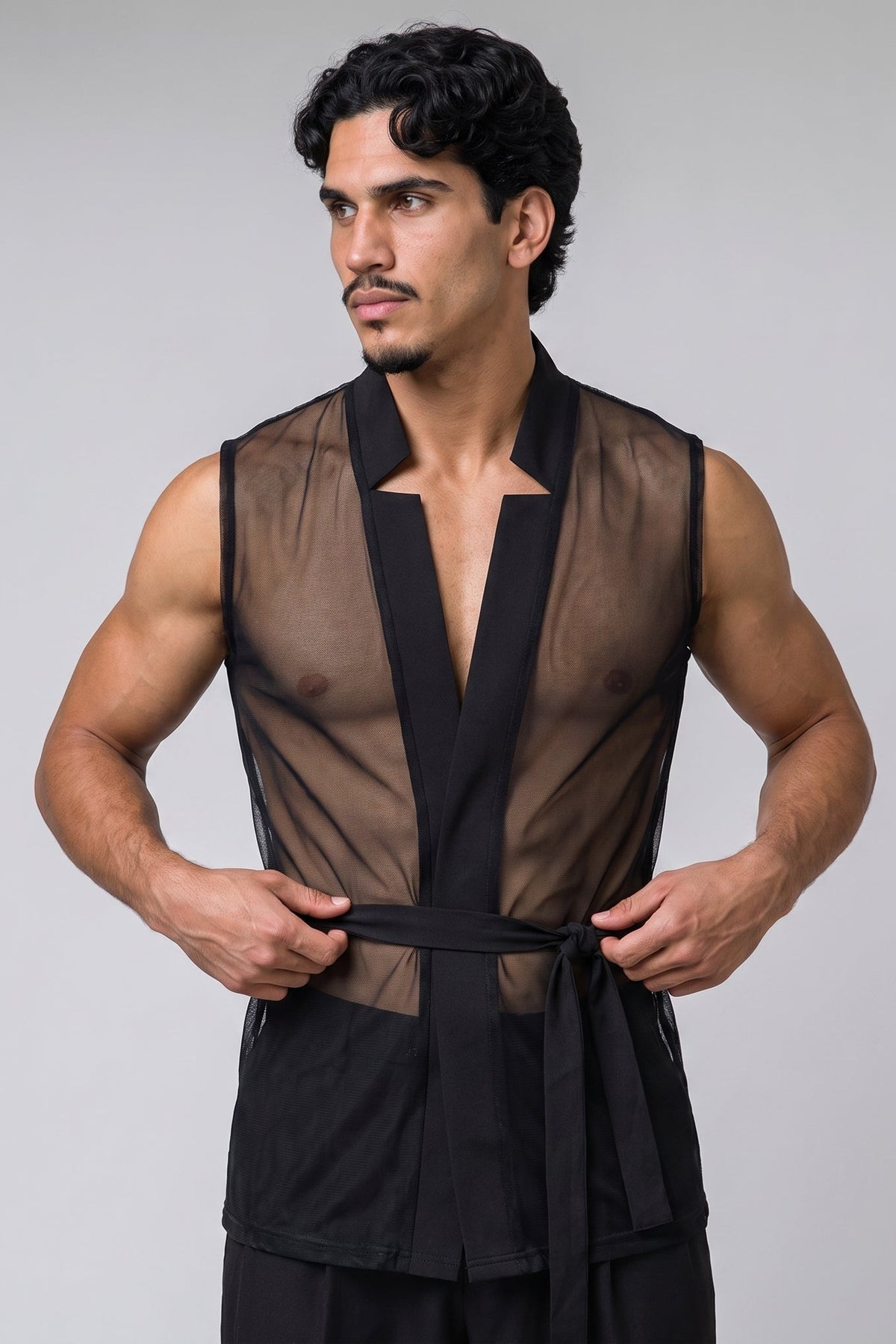 A muscular man wearing a black sheer mesh vest with a black waist tie, posing against a solid gray studio background.