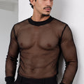 A man wearing a black long-sleeve fishnet mesh shirt and black trousers, standing against a white wall.