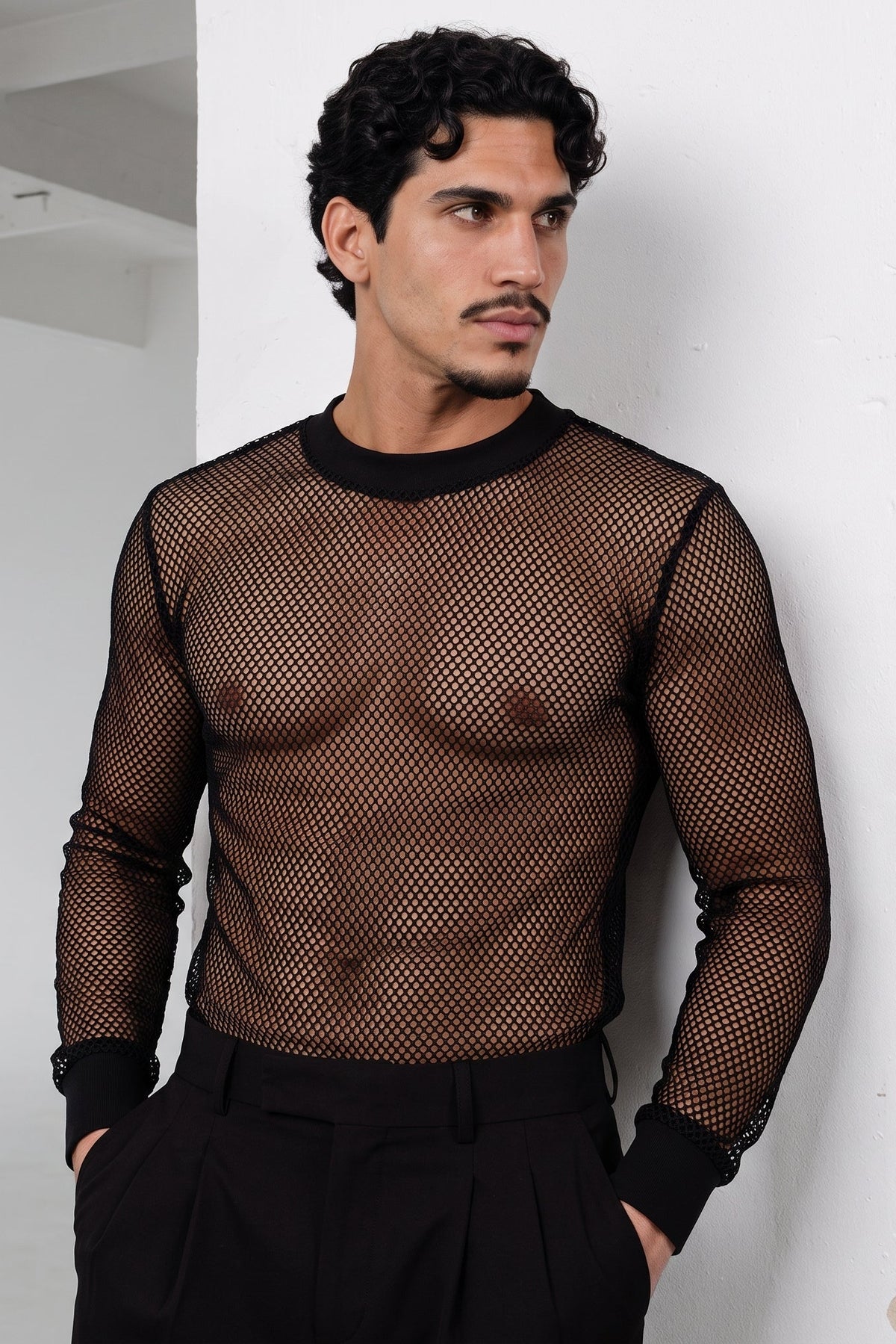A man wearing a black long-sleeve fishnet mesh shirt and black trousers, standing against a white wall.