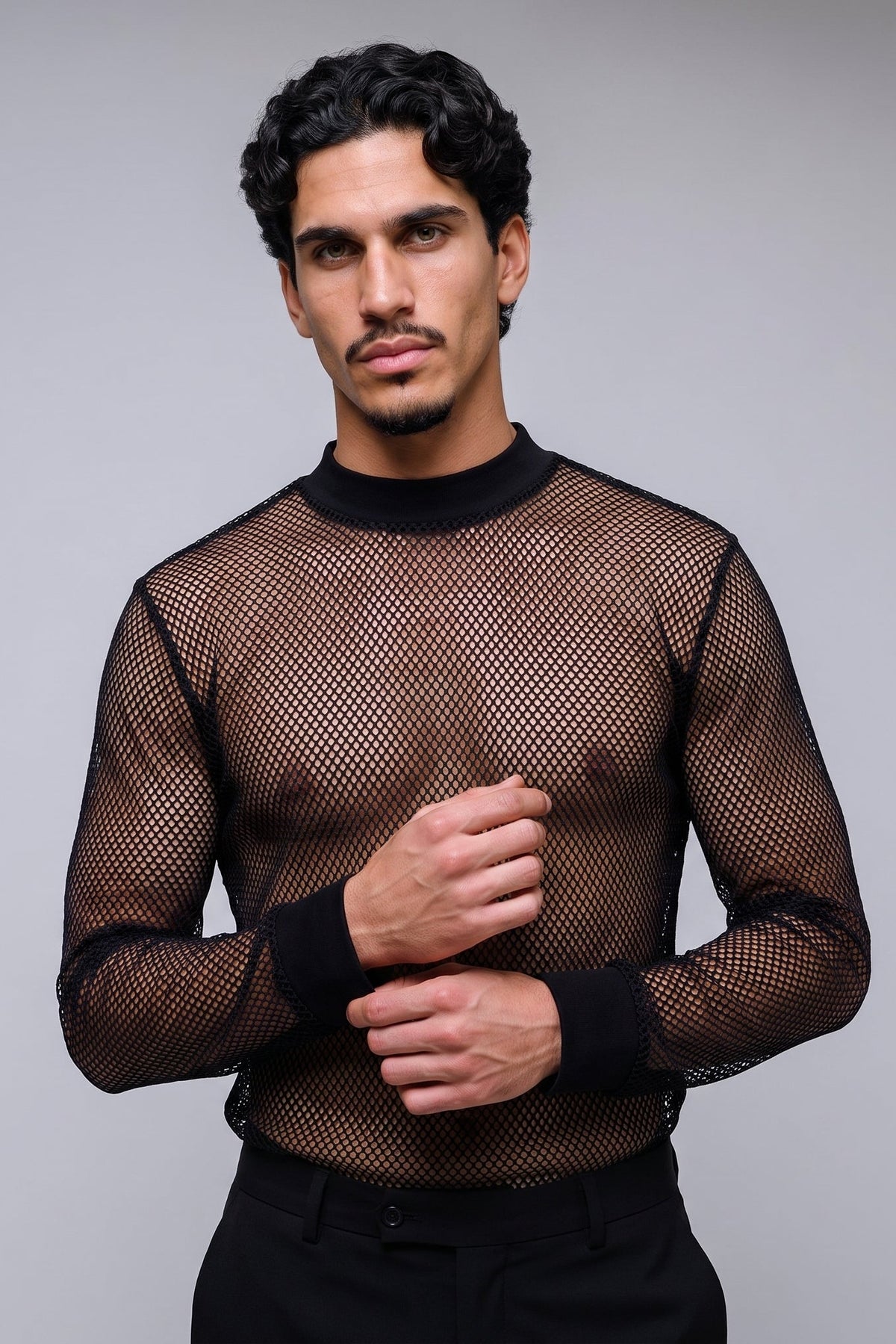 A man wearing a black long-sleeve mesh mock-neck shirt with black cuffs, standing against a solid gray studio background.