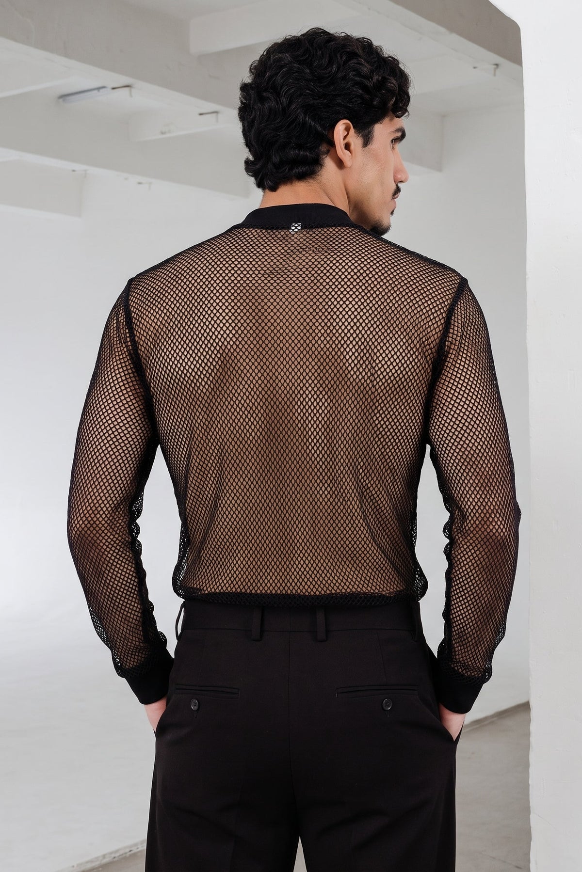 A man viewed from behind wearing a black long-sleeve mesh shirt and black trousers in a minimalist white room.