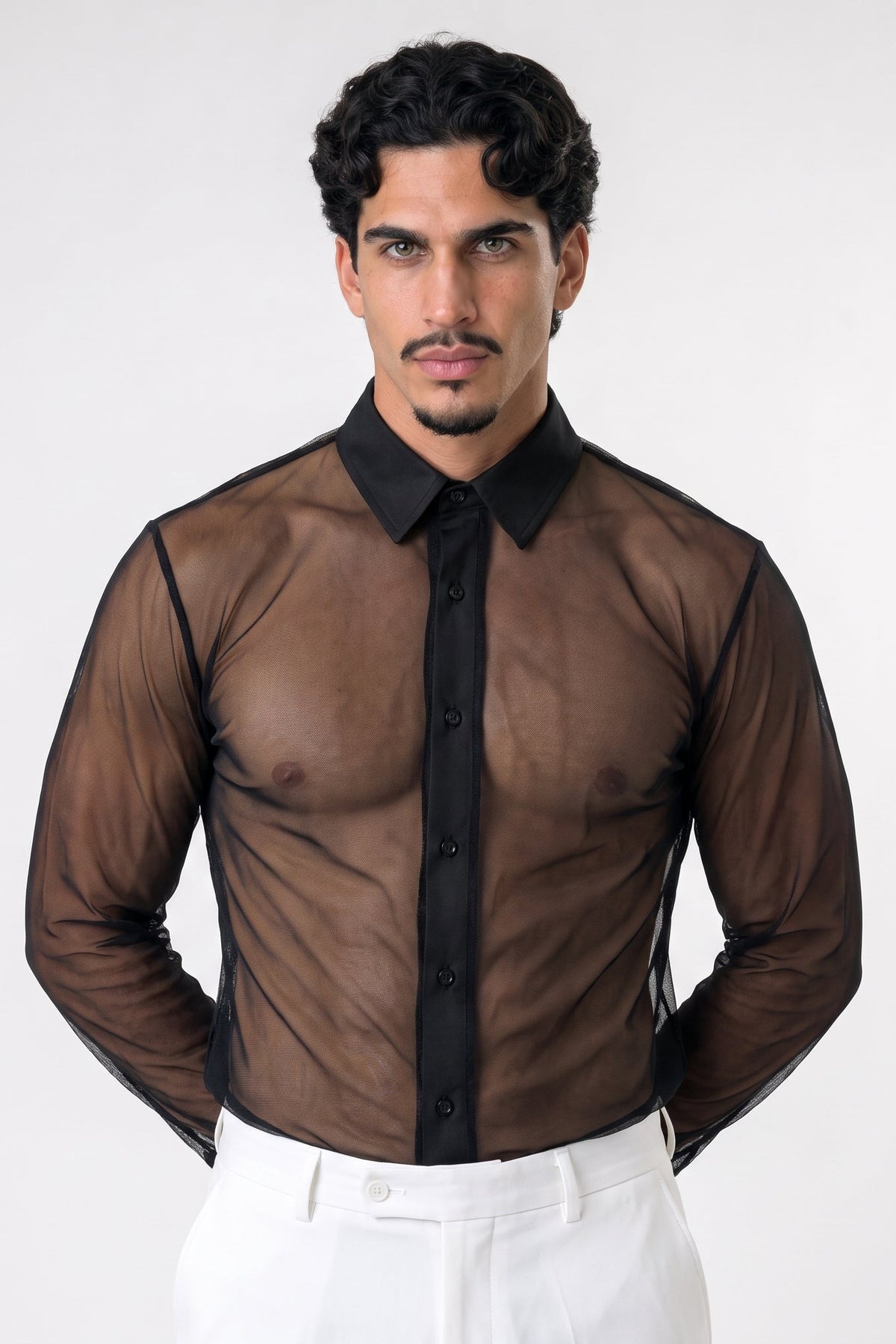 A man wearing a sheer brown long-sleeve button-up shirt with a black collar and black button placket, paired with white trousers, photographed against a light gray studio background.