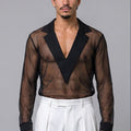 A man stands barefoot against a light gray studio background wearing a sheer black long-sleeve V-neck shirt with solid black lapels and white pleated trousers.