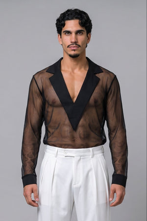 A man stands barefoot against a light gray studio background wearing a sheer black long-sleeve V-neck shirt with solid black lapels and white pleated trousers.