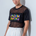 A male model wearing a black short-sleeve mesh crop top with a colorful 'PADAM' graphic on the chest, paired with white trousers, standing against a light gray wall.