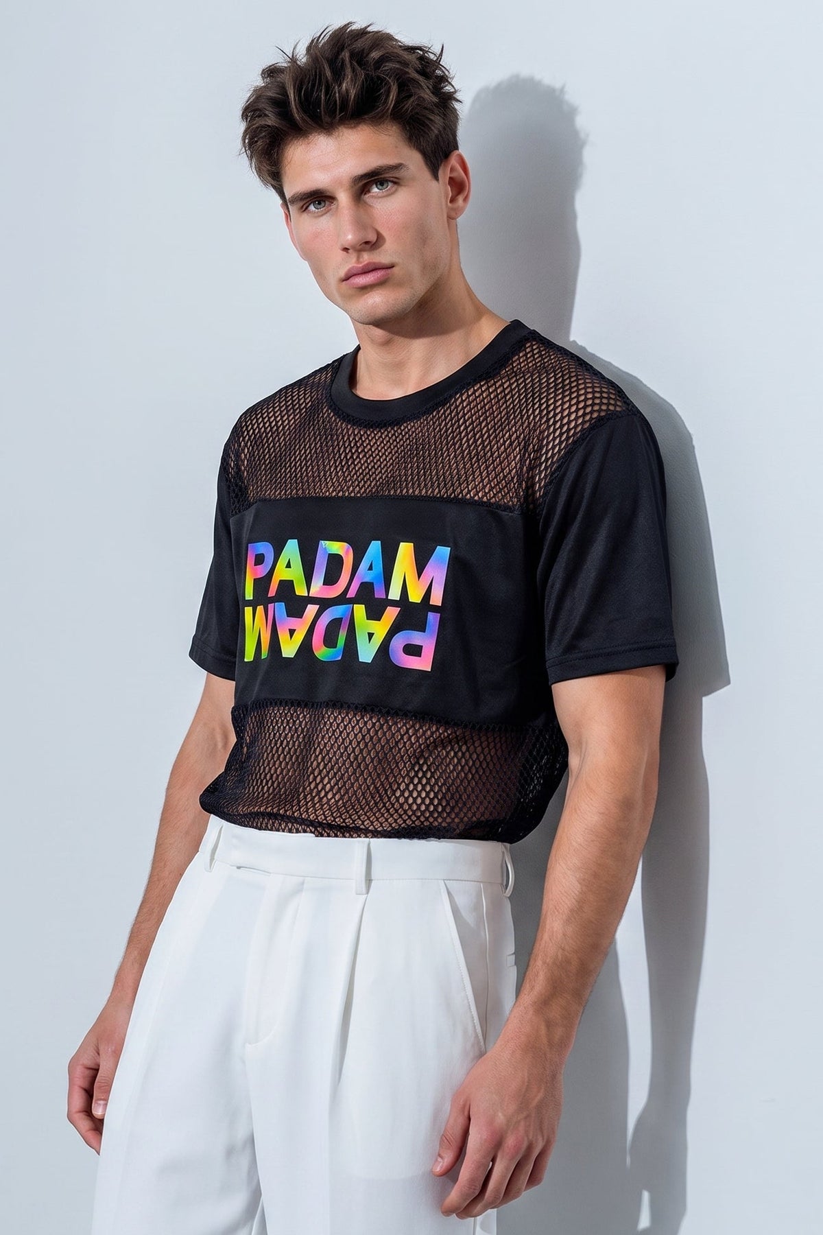 A male model wearing a black short-sleeve mesh crop top with a colorful 'PADAM' graphic on the chest, paired with white trousers, standing against a light gray wall.
