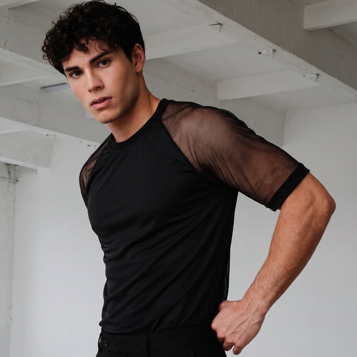 A man wearing a black short-sleeve athletic shirt with sheer mesh sleeves, posed in a minimalist white studio setting.