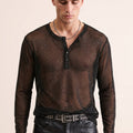 A man wearing a sheer black long-sleeve henley shirt with subtle glittery speckles, styled with black leather pants and a studded belt against a solid light beige background.