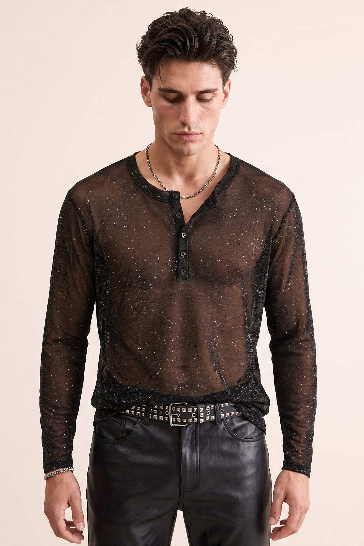 A man wearing a sheer black long-sleeve henley shirt with subtle glittery speckles, styled with black leather pants and a studded belt against a solid light beige background.
