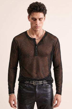 A man wearing a sheer black long-sleeve henley shirt with subtle glittery speckles, styled with black leather pants and a studded belt against a solid light beige background.