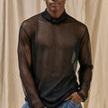 A man wearing a sheer black long-sleeve mock neck t-shirt and blue jeans, standing against a beige draped fabric backdrop.