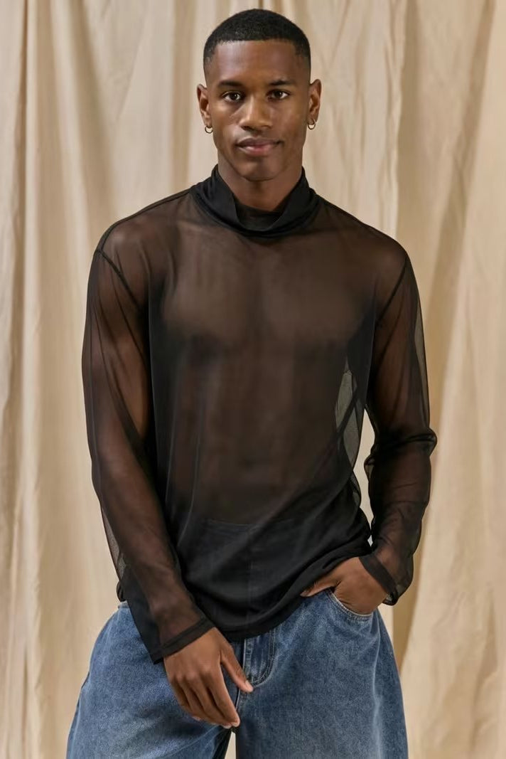 A man wearing a sheer black long-sleeve mock neck t-shirt and blue jeans, standing against a beige draped fabric backdrop.