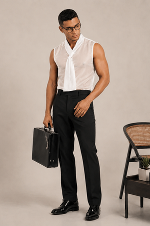 A man wearing a sleeveless white shirt with a tie-neck detail and black trousers, holding a black briefcase in a studio setting with a chair and small table.