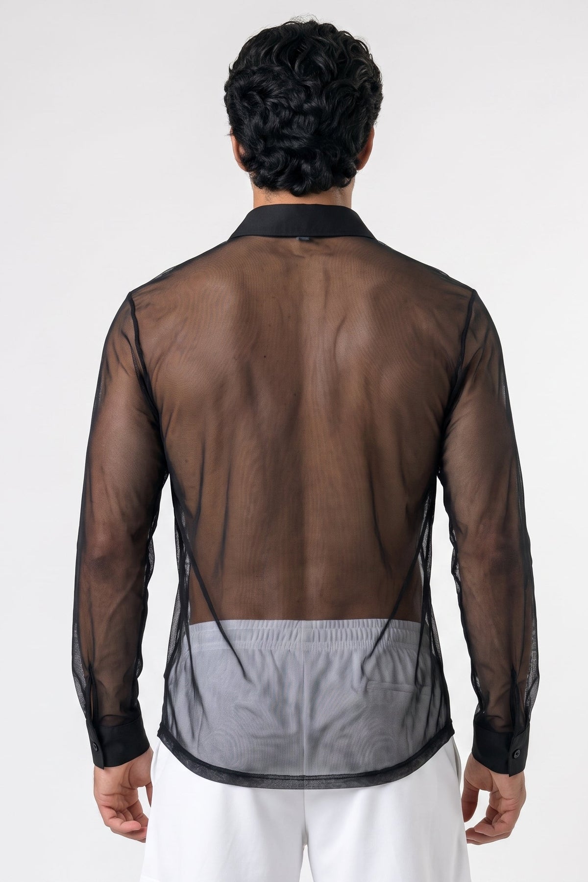 A man viewed from the back wearing a sheer black long-sleeve shirt over white pants, against a plain light gray studio background.
