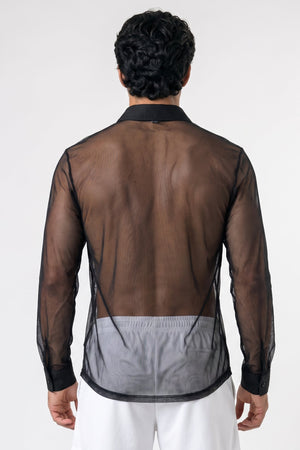 A man viewed from the back wearing a sheer black long-sleeve shirt over white pants, against a plain light gray studio background.