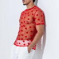 A man wearing a sheer red short-sleeve mesh top with a red heart pattern, paired with white trousers, leaning against a white wall.