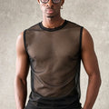 A man wearing a sheer black mesh sleeveless top over a brown undershirt, paired with brown trousers, standing against a textured gray background.