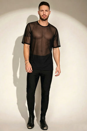 A man modeling a sheer black short-sleeve mesh top over a black t-shirt, paired with black pants and black boots, standing against a warm off-white studio background with a spotlight effect.