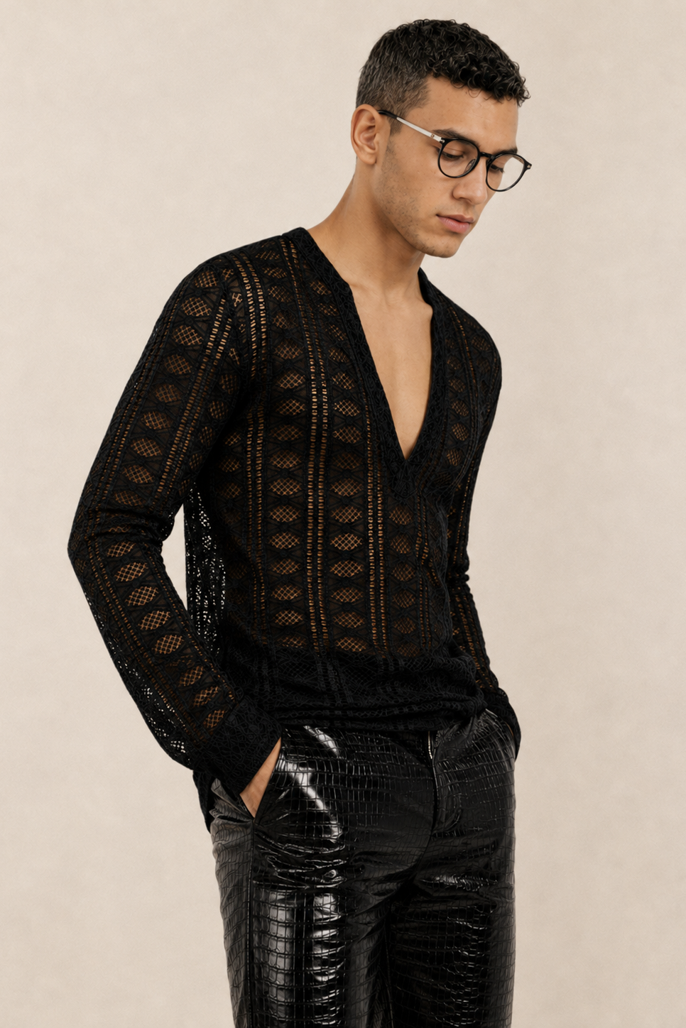 A man wearing a black open-knit long-sleeve V-neck sweater and glossy black pants, photographed against a plain beige studio background.