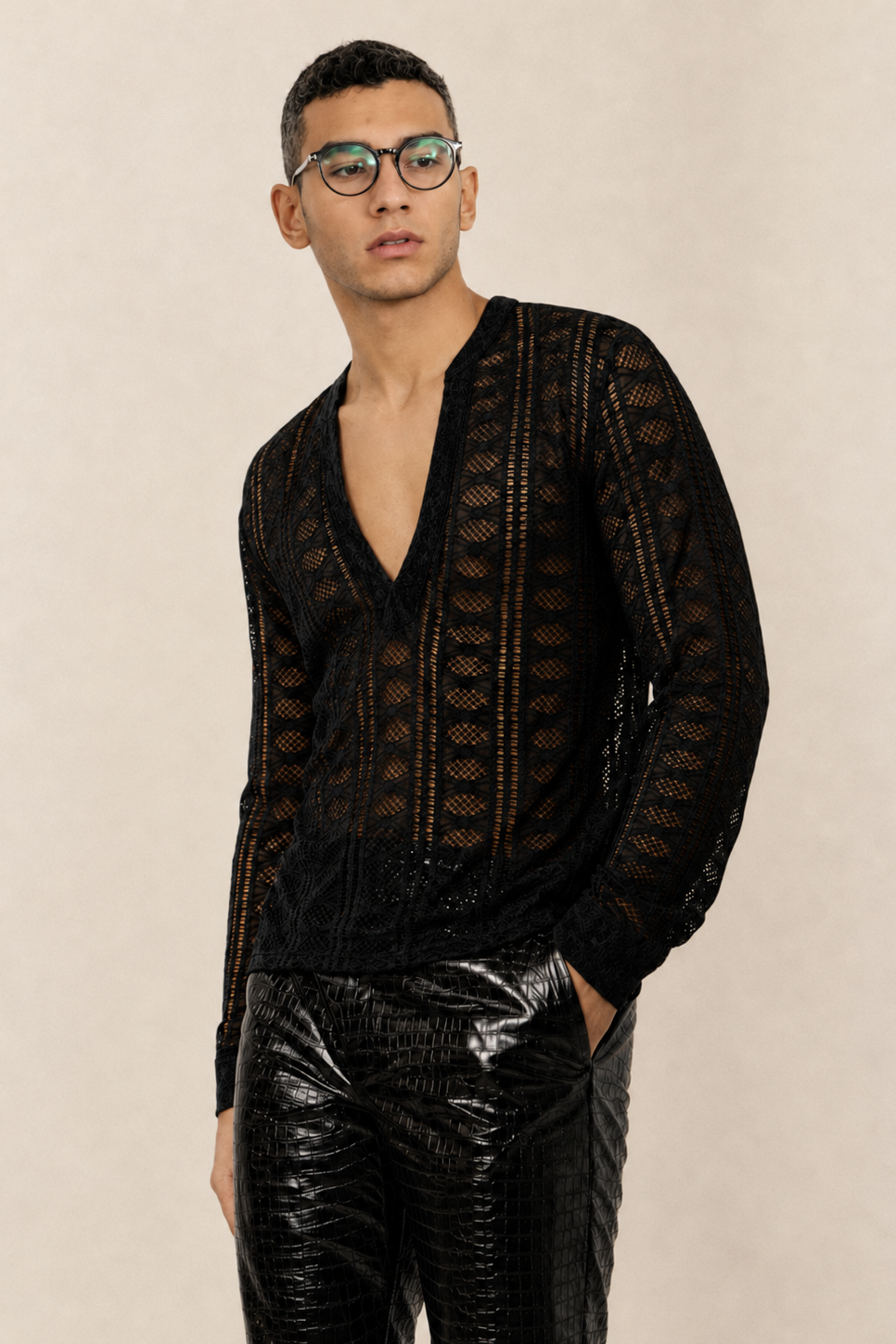 A man wearing a black open-knit long-sleeve cardigan with a deep V-neck and glossy black pants, standing against a textured beige studio background.