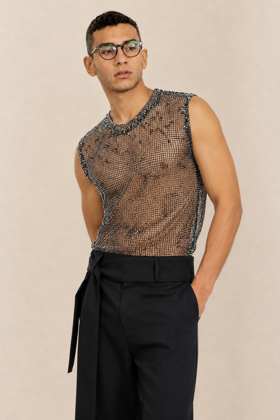 A man wearing a sleeveless black mesh top with a snakeskin pattern and black trousers, standing against a plain beige background.