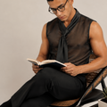 A man wearing a sheer black sleeveless shirt and black trousers sits on a woven chair, reading an open book in a minimalist studio setting.
