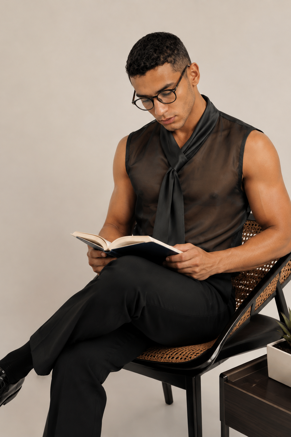 A man wearing a sheer black sleeveless shirt and black trousers sits on a woven chair, reading an open book in a minimalist studio setting.