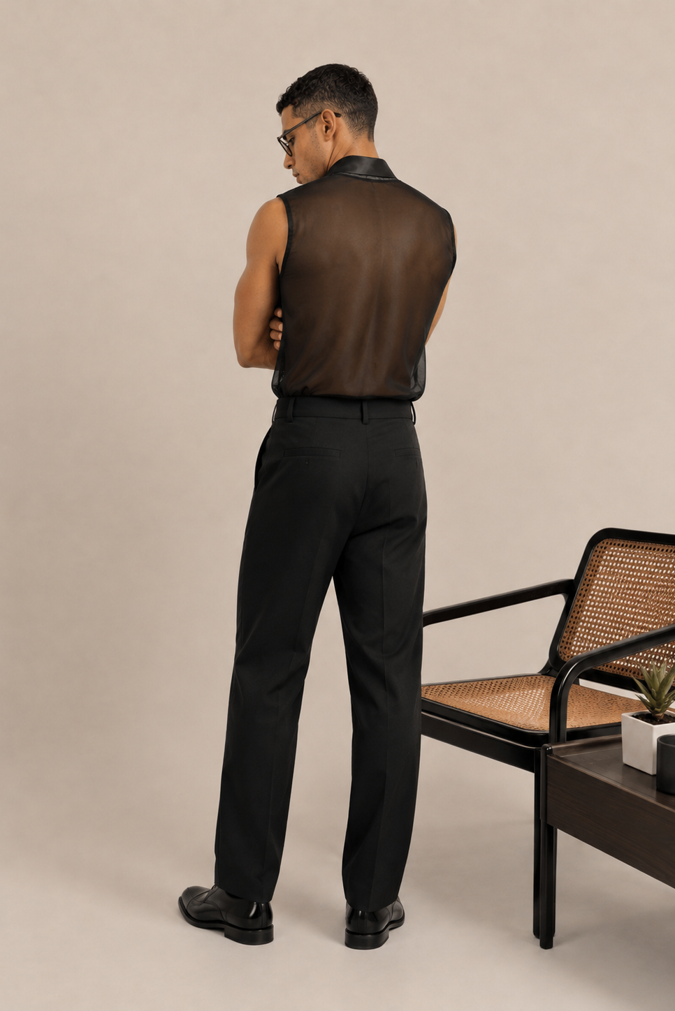 A man stands with his back to the camera wearing a sleeveless black mesh shirt, black trousers, and black dress shoes in a studio setting with a woven chair and side table.