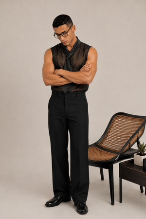 A man wearing a black sheer sleeveless shirt with a tie-neck detail, black trousers, and black dress shoes, standing with arms crossed next to a woven-back chair in a studio setting.
