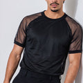 A man wearing a black short-sleeve athletic mesh shirt with sheer shoulder panels and black trousers, standing against a light gray wall.
