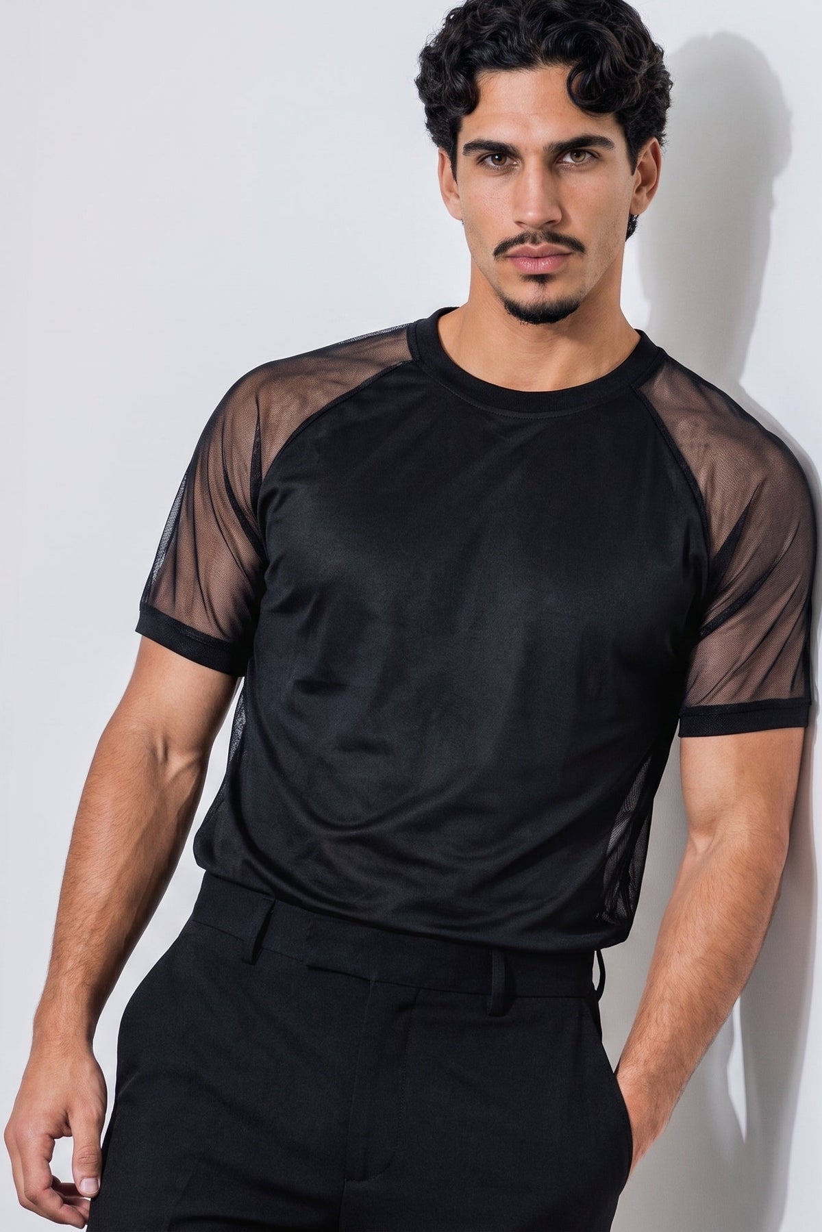 A man wearing a black short-sleeve athletic mesh shirt with sheer shoulder panels and black trousers, standing against a light gray wall.