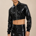 A man wearing a shiny black cropped zip-up jacket and matching high-waisted patent pants, photographed against a plain beige studio background.
