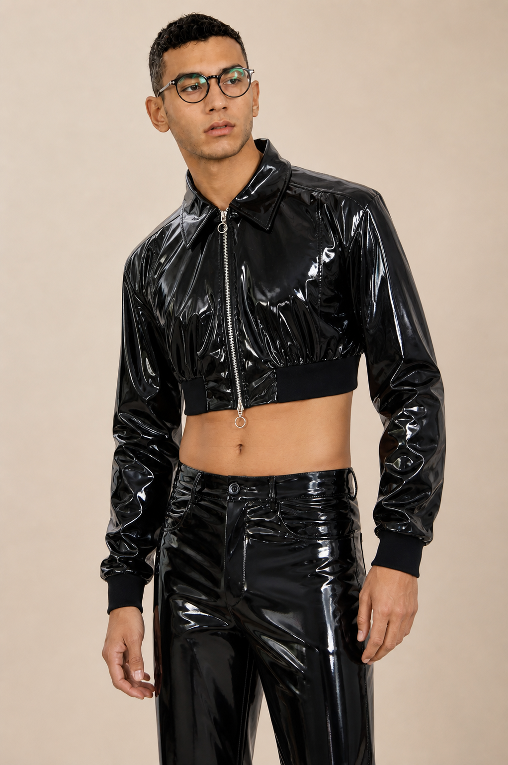 A man wearing a shiny black cropped zip-up jacket and matching high-waisted patent pants, photographed against a plain beige studio background.