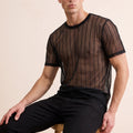 A man wearing a sheer black short-sleeve mesh t-shirt with vertical stripes and black pants, sitting on a wooden stool against a solid beige background.